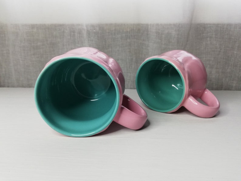 May include: Two ceramic mugs with pink exteriors and teal interiors. The mugs have a rounded shape and a handle on the side.