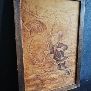 May include: A framed wood-burned artwork depicting a girl swinging on a rope swing in a forest setting. The artwork is in a brown wooden frame with a light brown background. The girl is wearing a dark brown dress and a white hat. The artwork is signed "Rune" in the bottom right corner.