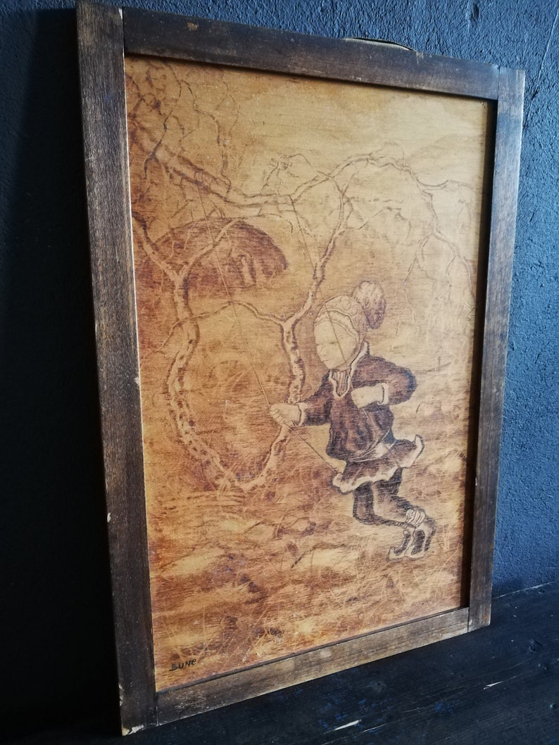 May include: A framed wood-burned artwork depicting a girl swinging on a rope swing in a forest setting. The artwork is in a brown wooden frame with a light brown background. The girl is wearing a dark brown dress and a white hat. The artwork is signed "Rune" in the bottom right corner.