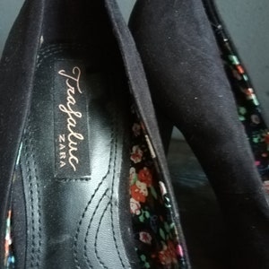 Op de afbeelding: Een paar zwarte su&egrave;de hoge hakken met een bloemenprint voering. De schoenen hebben een label met de tekst "Trafaluc ZARA".