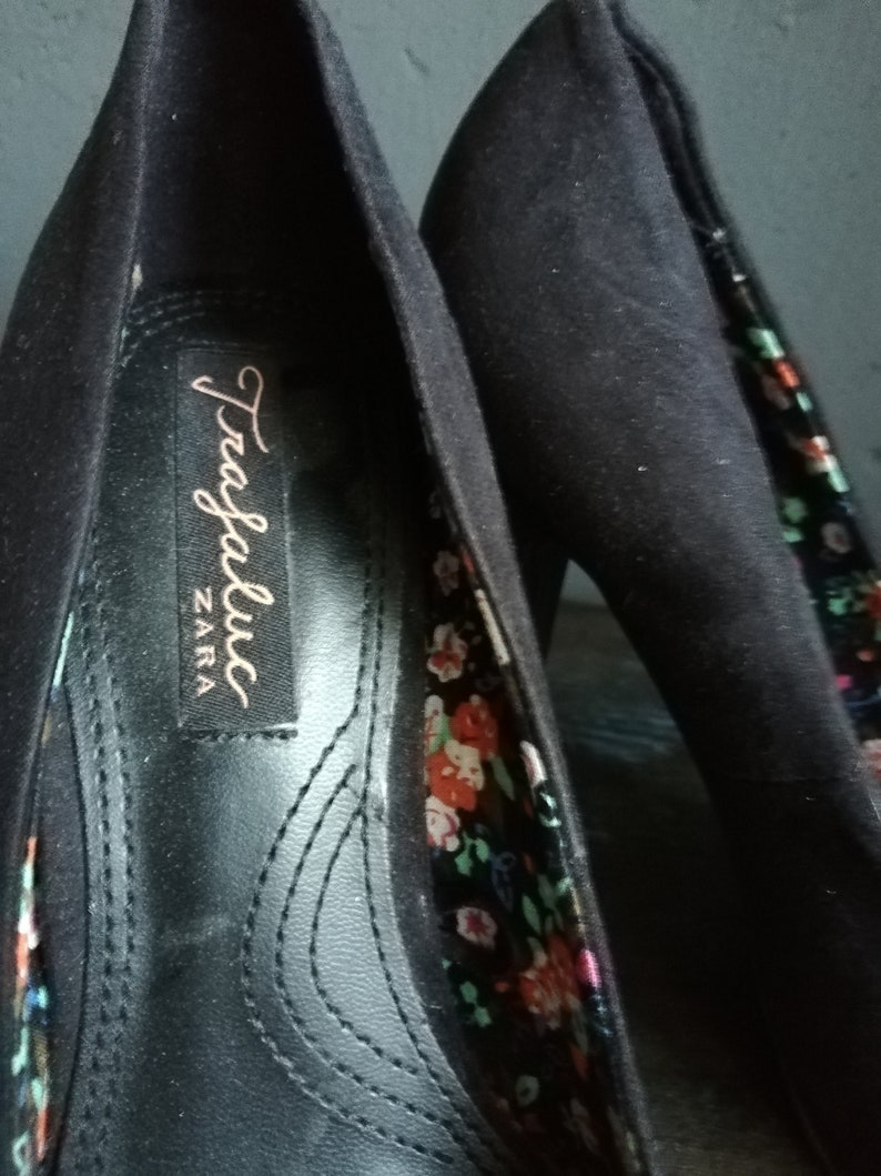 Op de afbeelding: Een paar zwarte su&egrave;de hoge hakken met een bloemenprint voering. De schoenen hebben een label met de tekst "Trafaluc ZARA".