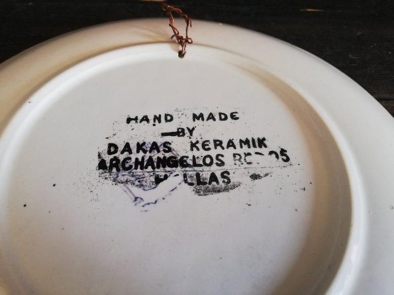 May include: White ceramic plate with a black stamp that reads "HAND MADE - BY - DAKAS KERAMIK ARCHANGELOS RE-05 LLAS".