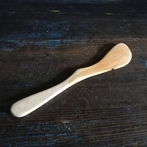 May include: A wooden butter knife with a light brown handle and a white blade. The knife is lying on a dark brown wooden surface.