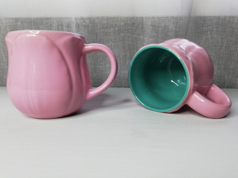 May include: Two ceramic mugs, one pink with a light green interior and one light green with a pink interior. Both mugs have a rounded shape and a handle.