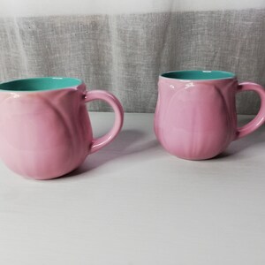 May include: Two pink ceramic mugs with a turquoise interior. The mugs are shaped like tulips and have a rounded handle.