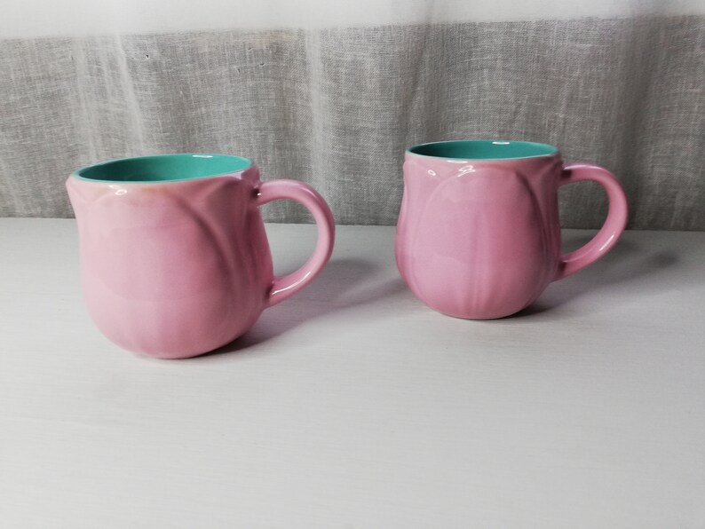 May include: Two pink ceramic mugs with a turquoise interior. The mugs are shaped like tulips and have a rounded handle.