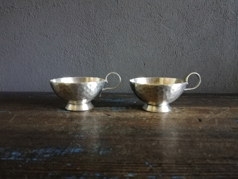 May include: Two silver hammered metal cups with handles. The cups are sitting on a dark wooden surface.