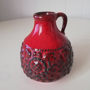 May include: A red ceramic vase with a handle and an embossed floral design.