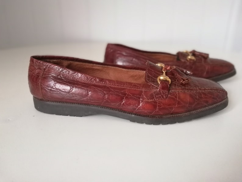Puede incluir: Un par de mocasines de cuero marr&oacute;n con un detalle de borla y herrajes dorados. Los mocasines tienen un patr&oacute;n de piel de cocodrilo.