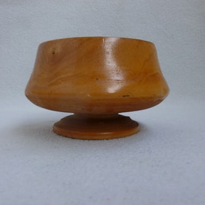 May include: A small, wooden bowl with a flared lip and a pedestal base. The bowl is made of light brown wood and has a smooth, polished finish.