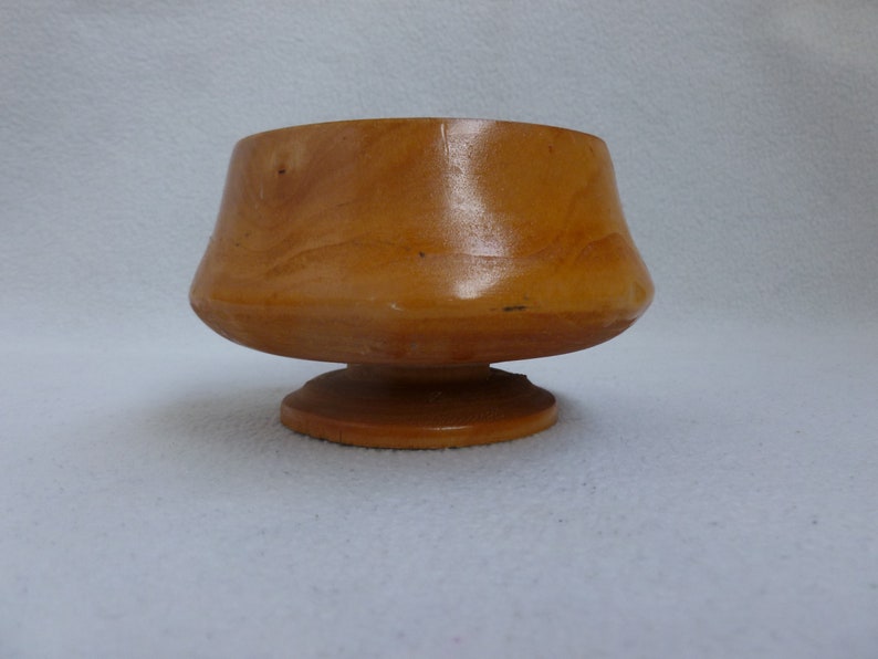 May include: A small, wooden bowl with a flared lip and a pedestal base. The bowl is made of light brown wood and has a smooth, polished finish.