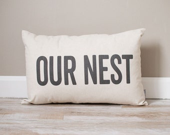 May include: A white rectangular pillow with black text that reads "OUR NEST".