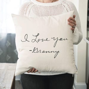 May include: Cream-colored square pillow with the handwritten text "I Love you -Granny" in black script. The pillow is held by a person wearing a white sweater and dark pants. The pillow is a decorative home item.