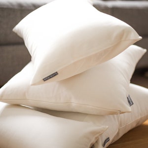 May include: A stack of four off-white square pillows. Each pillow has a tag on the side. The pillows are stacked on top of each other, with the top pillow angled towards the viewer. The background is blurred, showing a couch and a wooden surface.