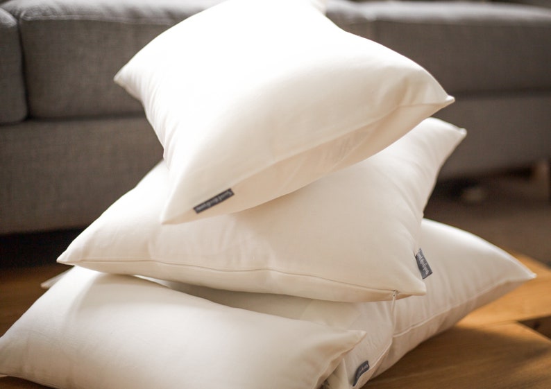 May include: A stack of four off-white square pillows. Each pillow has a tag on the side. The pillows are stacked on top of each other, with the top pillow angled towards the viewer. The background is blurred, showing a couch and a wooden surface.