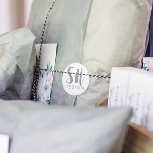May include: A gift box filled with tissue paper and wrapped with a black and white striped ribbon. A white circle sticker with the text "SH DESIGN" is attached to the tissue paper.