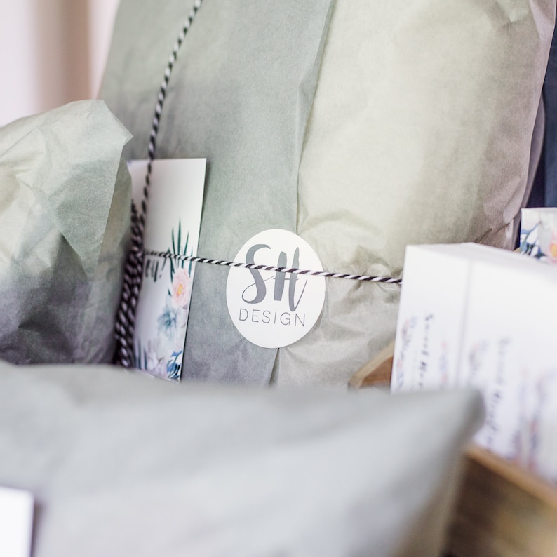May include: A gift box filled with tissue paper and wrapped with a black and white striped ribbon. A white circle sticker with the text "SH DESIGN" is attached to the tissue paper.