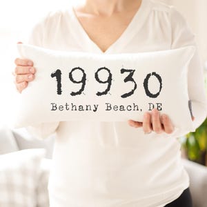 May include: A white rectangular cushion featuring the text "19930 Bethany Beach, DE" in black. The cushion is held by a person wearing a white long-sleeved top.