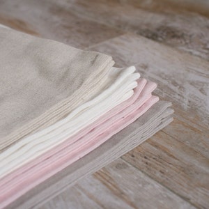 May include: A stack of folded linen napkins in various shades of beige, white, and pink. The napkins are arranged in a graduated color scheme, with the lightest shade at the top and the darkest shade at the bottom.