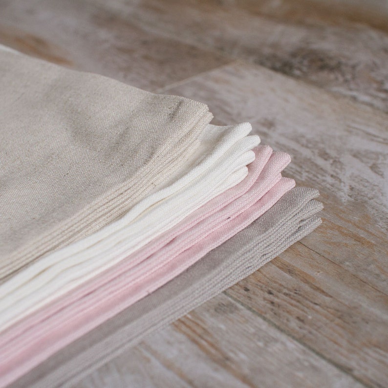 May include: A stack of folded linen napkins in various shades of beige, white, and pink. The napkins are arranged in a graduated color scheme, with the lightest shade at the top and the darkest shade at the bottom.