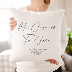 May include: A square, off-white pillow with the phrase "Mi Casa es Tu Casa" in black script. Below the phrase is the text "The Henderson's, Moseley, VA, Est. 2025". The pillow is held by a person.