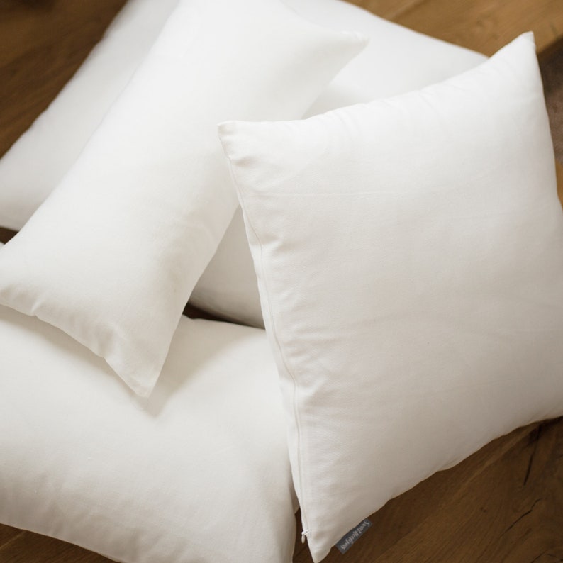 May include: Four white throw pillows arranged on a wooden surface. The pillows are made of a soft, smooth fabric and are all the same size.