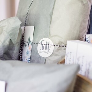 May include: Gift-wrapped package in light green tissue paper, tied with black and white twine. A white circular tag with "SH DESIGN" in gray script is attached. A floral card is visible.