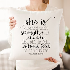 May include: White throw pillow with black text that reads "She is clothed with strength and dignity and she laughs without fear of the future Proverbs 31:25".