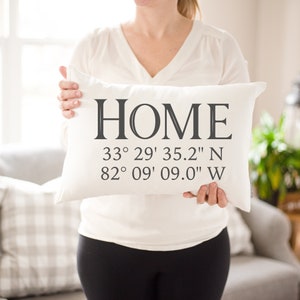 May include: A rectangular white pillow with the word "HOME" printed in large, dark letters. Below the word are coordinates: 33&deg; 29' 35.2" N and 82&deg; 09' 09.0" W. The pillow is held by a person.