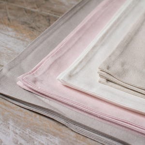 May include: A stack of four linen napkins in different shades of beige, pink, and white. The napkins are folded and arranged in a graduated color scheme, with the darkest shade at the bottom and the lightest shade at the top.
