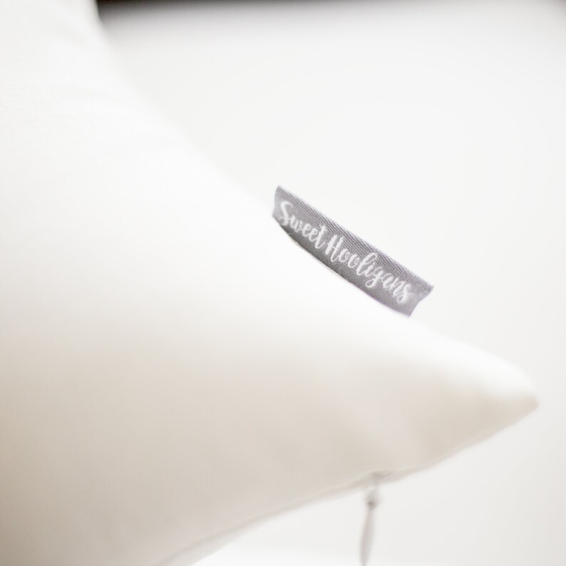 May include: A white pillow with a gray tag that reads "Sweet Hooligans" in cursive. The pillow is a solid white color and appears to be made of a soft material. The tag is attached to the side of the pillow.