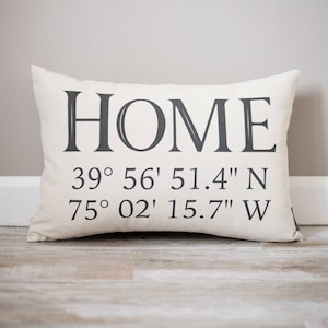 May include: Rectangular off-white pillow with the word "HOME" printed in dark gray. Below the word are coordinates: 39&deg; 56' 51.4" N and 75&deg; 02' 15.7" W. The pillow is on a light wood floor.