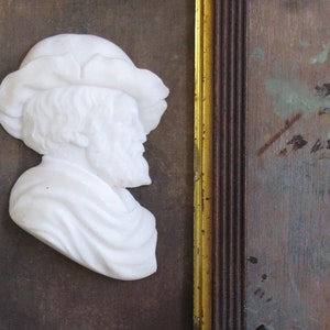 May include: A white marble bust of a person wearing a hat, framed in a gold and brown frame.