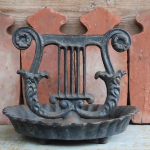 May include: A cast iron decorative dish in the shape of a lyre. The dish is black with ornate scrollwork and a shallow bowl. The background features red-brown tiles and a wooden surface.