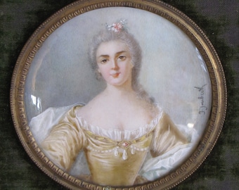 May include: A miniature portrait of a woman in a gold dress with a white lace collar. She is wearing a flower crown and has her hair pulled back. The portrait is framed in a gold frame and signed "J. Jacob" in the lower right corner.