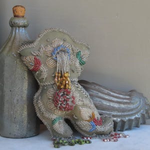 May include: A decorative, sage green, beaded boot-shaped pillow with floral and leaf designs. The pillow features red, blue, and yellow bead accents and a beaded fringe. A gray bottle with a wooden stopper and a decorative shell are in the background.