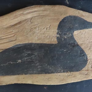 May include: A rustic, wooden wall art piece featuring a painted silhouette of a loon. The loon is depicted in black against a light brown, textured background. The artwork has a natural, organic shape and is likely handmade.