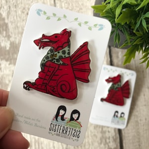 以下が含まれることがあります： 緑色のスカーフをつけた赤いドラゴンのピンが2つ。ピンは、「Hand made on the Shropshire/Welsh Borders」と「Sisteract212」の文字が書かれた白いカードの上に置かれています。