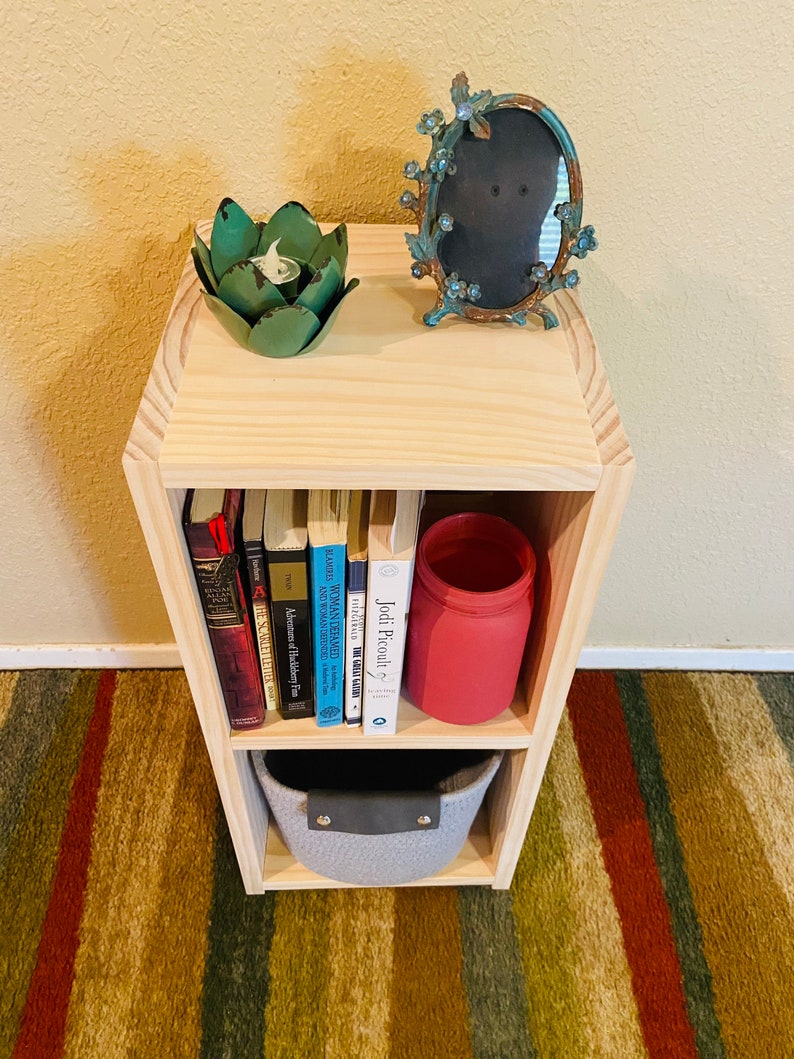 Op de afbeelding: Een houten boekenkast met twee planken, een grijze opbergmand, een rode pot en een aantal boeken. De boekenkast staat op een gestreept tapijt.