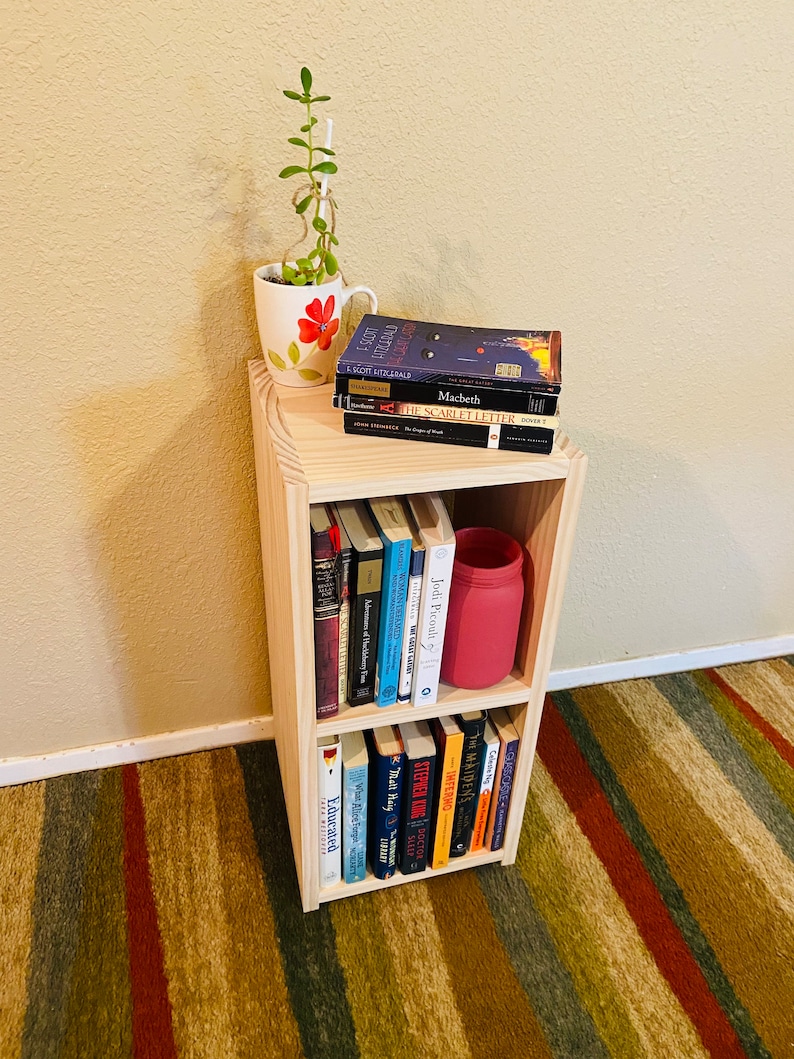 Op de afbeelding: Een houten boekenkast met twee planken, gevuld met boeken. Een rode pot en een mok met een bloemenpatroon staan op de bovenste plank. De boekenkast staat op een gestreept tapijt.