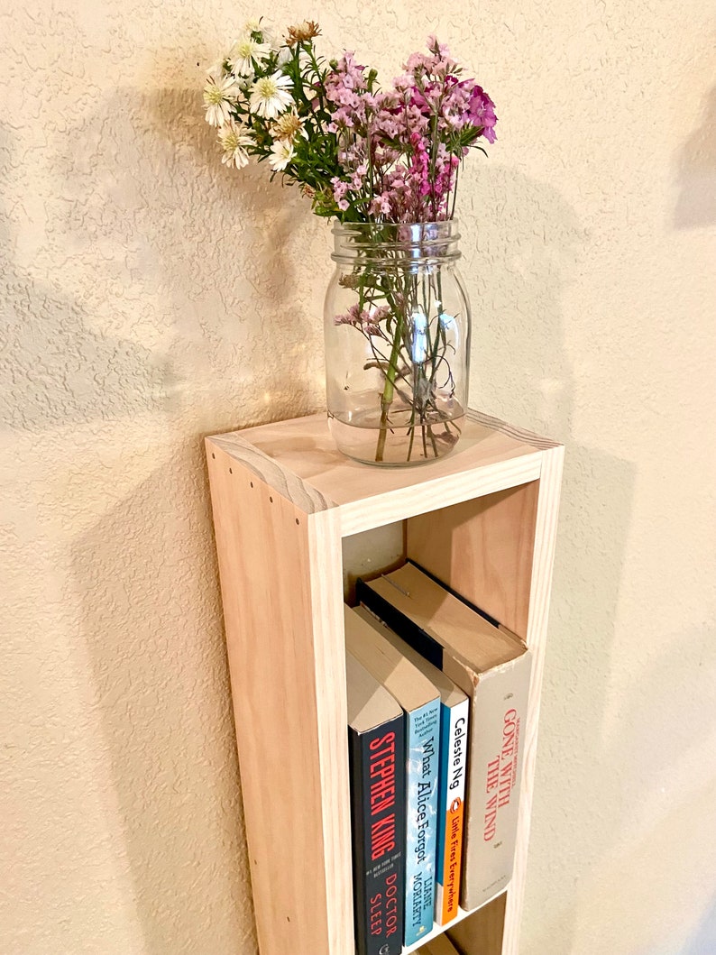 May include: A wooden bookshelf with three shelves, holding a variety of books. The bookshelf is made of light wood and has a simple design. A mason jar filled with pink and white flowers sits on top of the bookshelf.