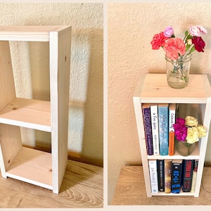 May include: A two-tier wooden bookshelf with a whitewash finish. The bookshelf is empty on the left side of the image and filled with books on the right side. The bookshelf is decorated with a vase of pink carnations.