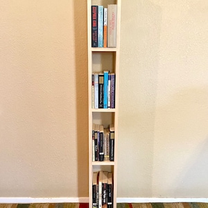 May include: A tall, narrow wooden bookshelf with four shelves, each filled with books. The bookshelf is made of light wood and has a simple, modern design.