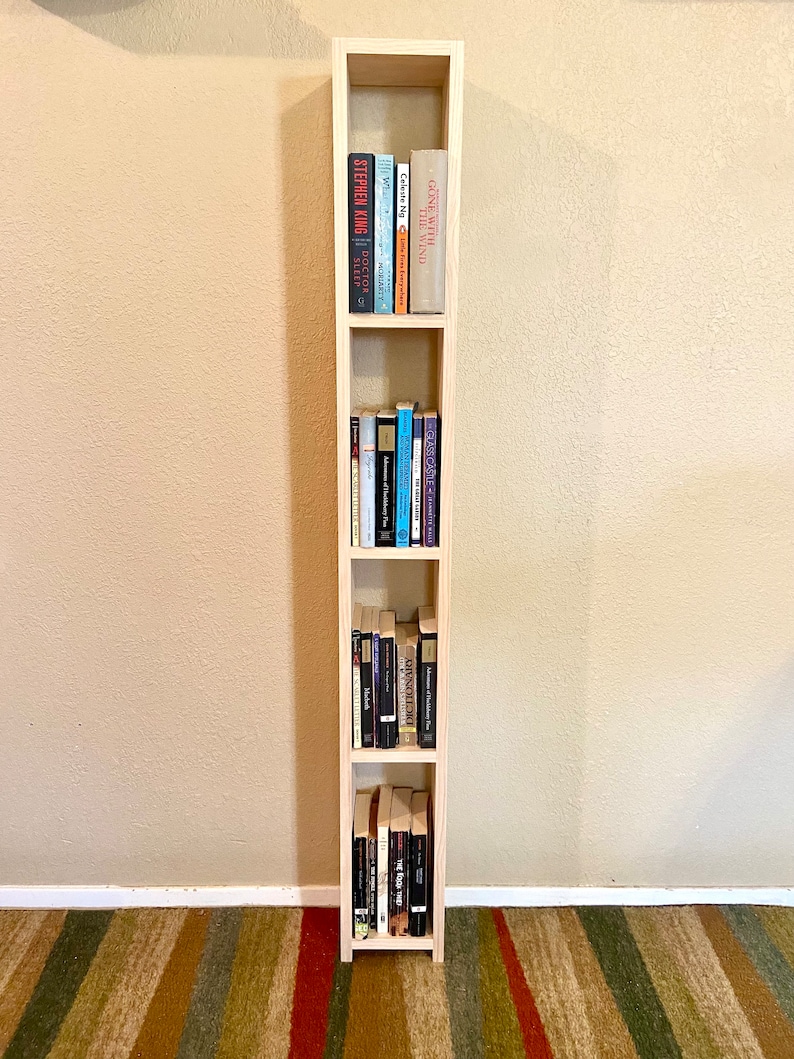 May include: A tall, narrow wooden bookshelf with four shelves, each filled with books. The bookshelf is made of light wood and has a simple, modern design.