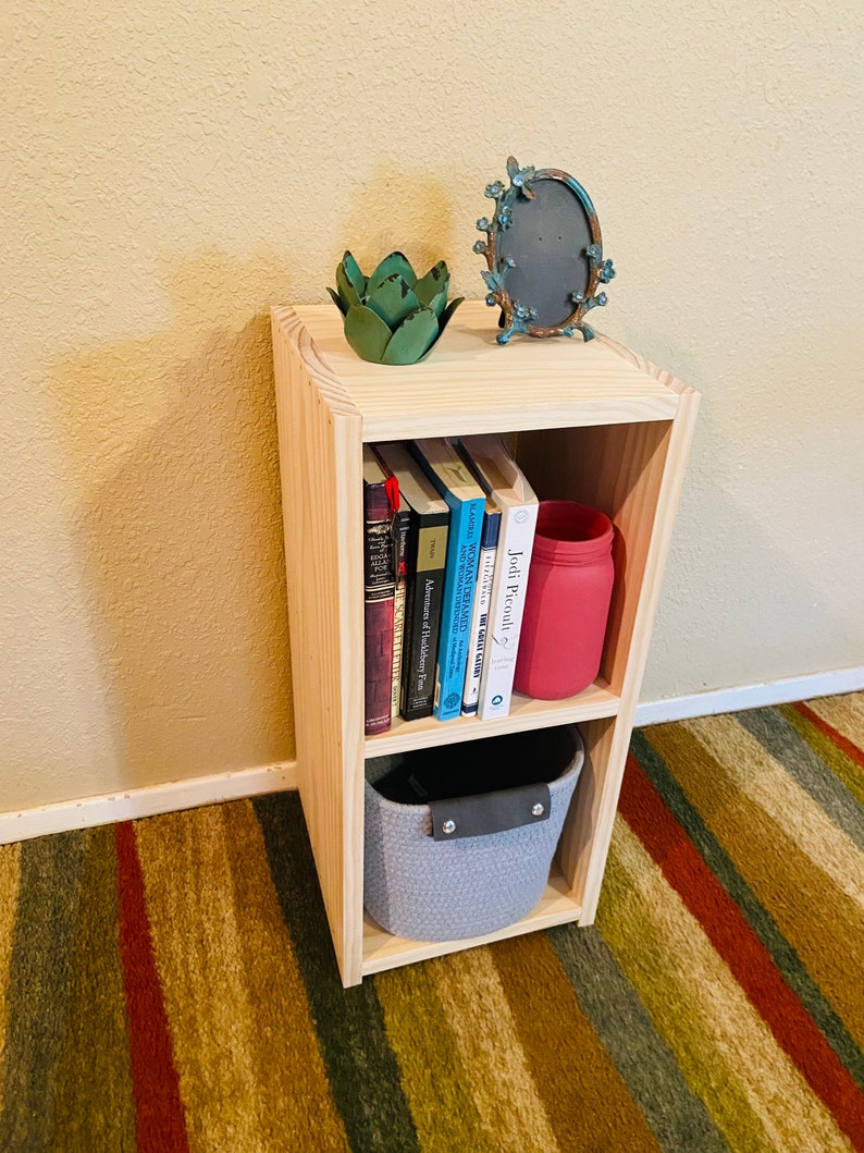 Op de afbeelding: Een houten plank met twee planken, een grijze geweven mand, een rode glazen pot en een verzameling boeken. De plank staat op een gestreept tapijt.