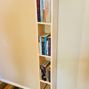 May include: A light wood bookshelf with four shelves, filled with various books. The bookshelf is standing in a corner of a room with a striped rug on the floor.