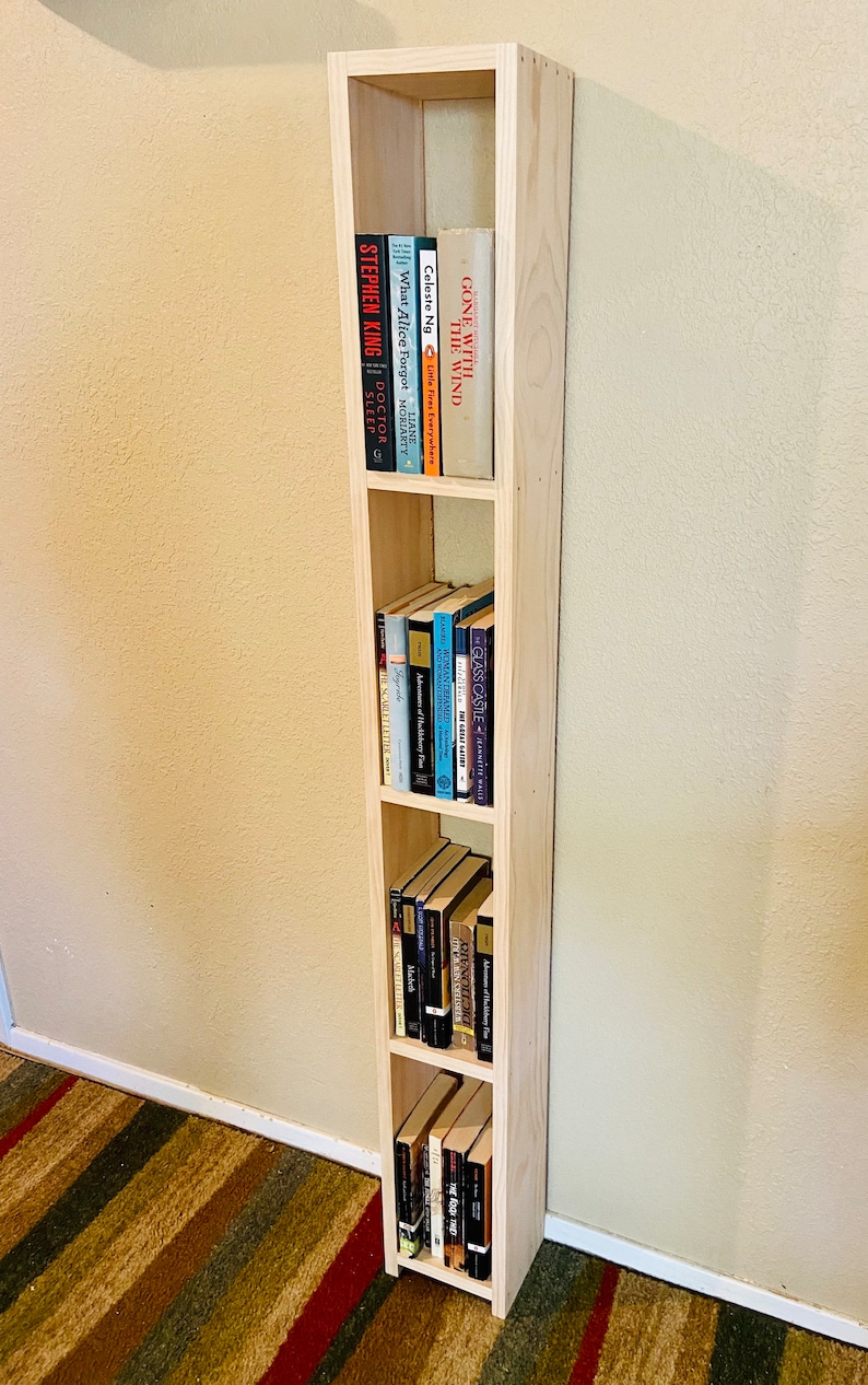 May include: A light wood bookshelf with four shelves, filled with various books. The bookshelf is standing in a corner of a room with a striped rug on the floor.