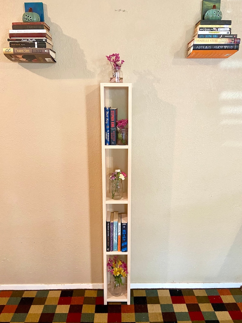 May include: A white wooden bookshelf with four shelves. The shelves are filled with books and small vases with flowers. The bookshelf is standing on a patterned rug.