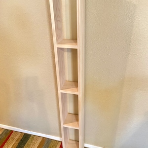 May include: A tall, unfinished wooden shelf unit with four open shelves. The shelf unit is standing against a light beige wall.