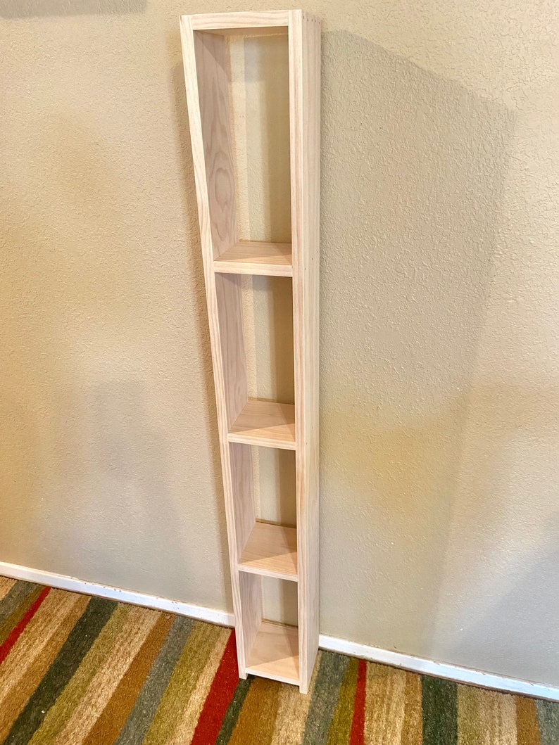 May include: A tall, unfinished wooden shelf unit with four open shelves. The shelf unit is standing against a light beige wall.